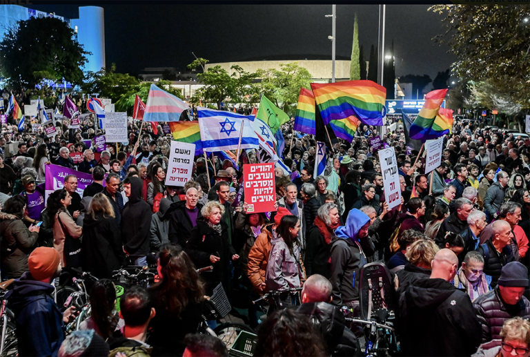 Una folla di Israeliani a una protesta contro il nuovo governo del Primo Ministro Benjamin Netanyahu, i manifestanti hanno bandiere di vario tipo (slogan in ebraico, bandiere della pace, di Israele, bandiera transgender)