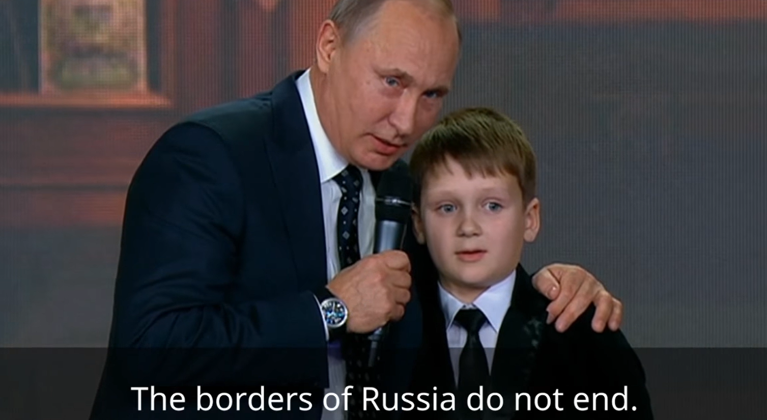 Vladimir Putin appoggia la mano sulla spalla di un bambino, il mento sulla testa, durante un evento pubblico, i sottotitoli in inglese recitano "i confini della Russia non hanno fine"