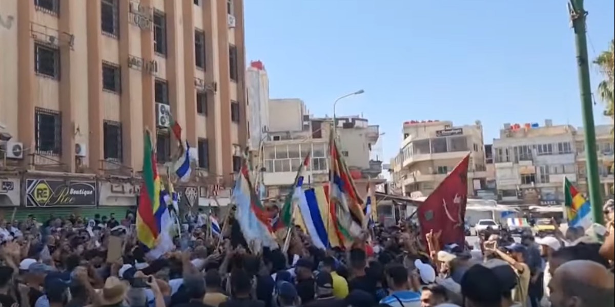 Una folla radunata protesta davanti a un edificio in Siria, issando varie bandiere.