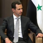 Il presidente della Siria Assad seduto su una poltrona, con alle spalle la bandiera siriana.