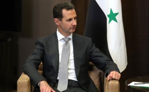 Il presidente della Siria Assad seduto su una poltrona, con alle spalle la bandiera siriana.