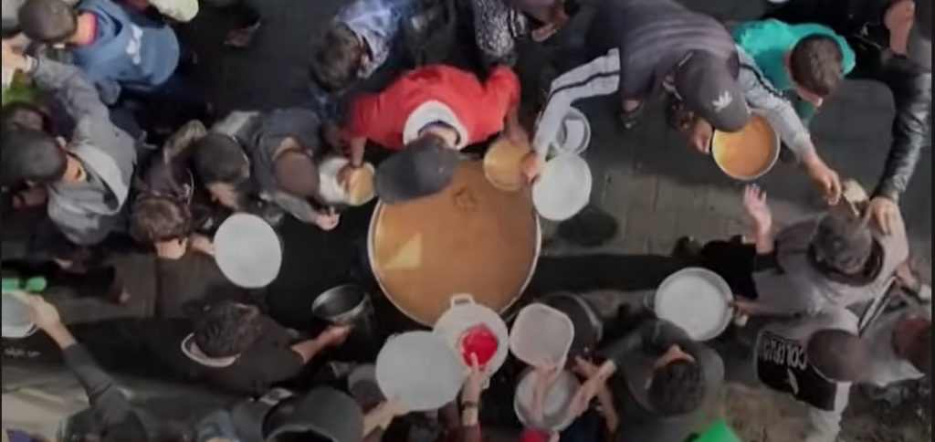 Immagine dall'alto che mostra un gruppo di civili attorno a una pentola per prendere una razione. Il rapporto di Human Rights Watch documenta come Israele abbia impedito l'accesso a cibo e acqua, affamando la popolazione civile a Gaza.