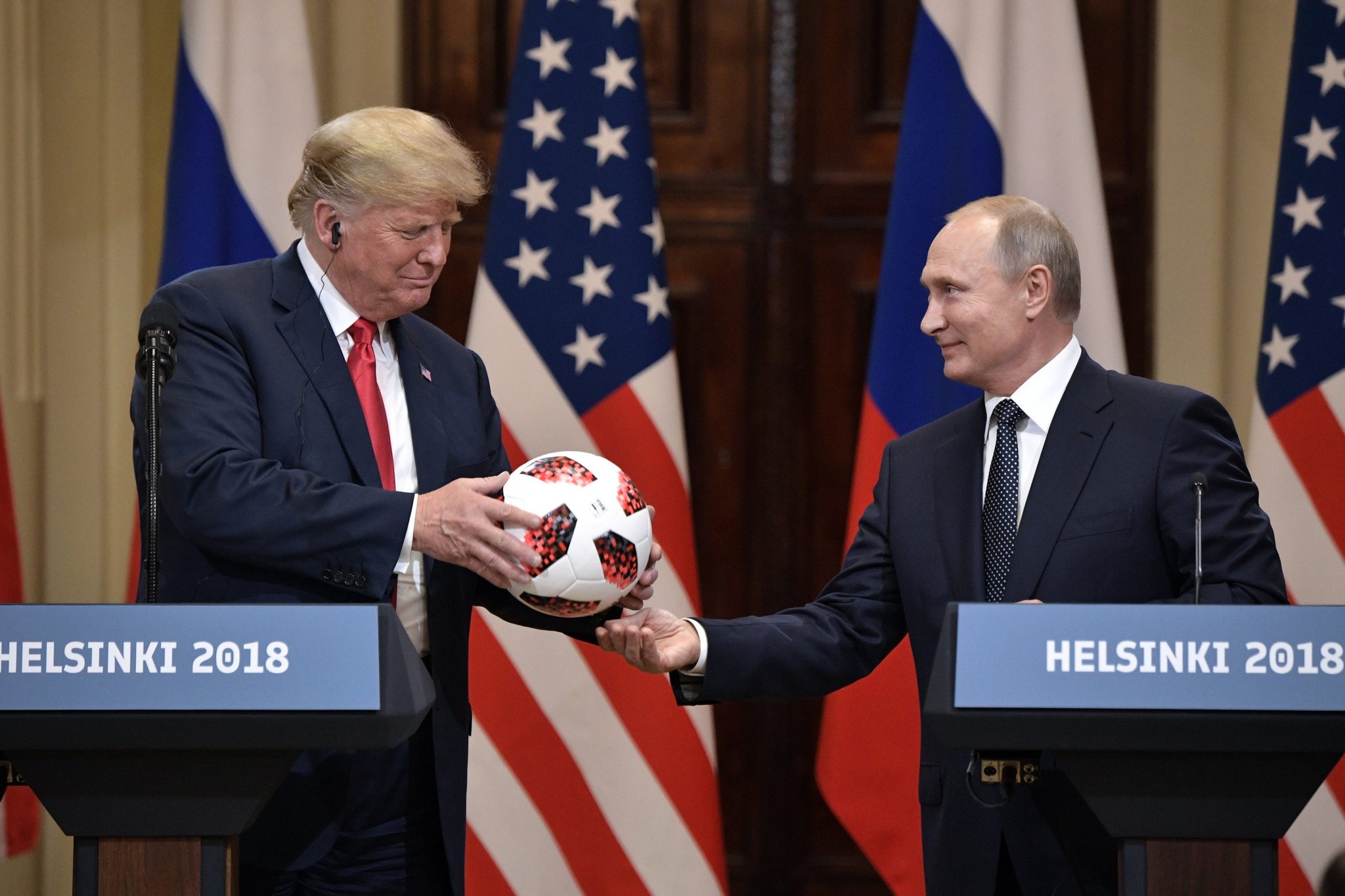 Trump e Putin dietro due leggii con scritto "Helsinki 2018". Trump osserva il pallone che ha tra le mani. Putin tende la mano sotto il pallone - non si capisce se gli abbia appena passato il pallone, o se Trump stia per passarlo a lui.