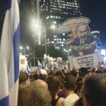 Una folla che protesta per le strade di Tel Aviv. Alcuni manifestanti reggono bandiere di Israele. Un cartello mostra una caricatura del premier Nethanyahu mentre impugna una valigia piena di banconote e ha alle spalle un fungo atomico