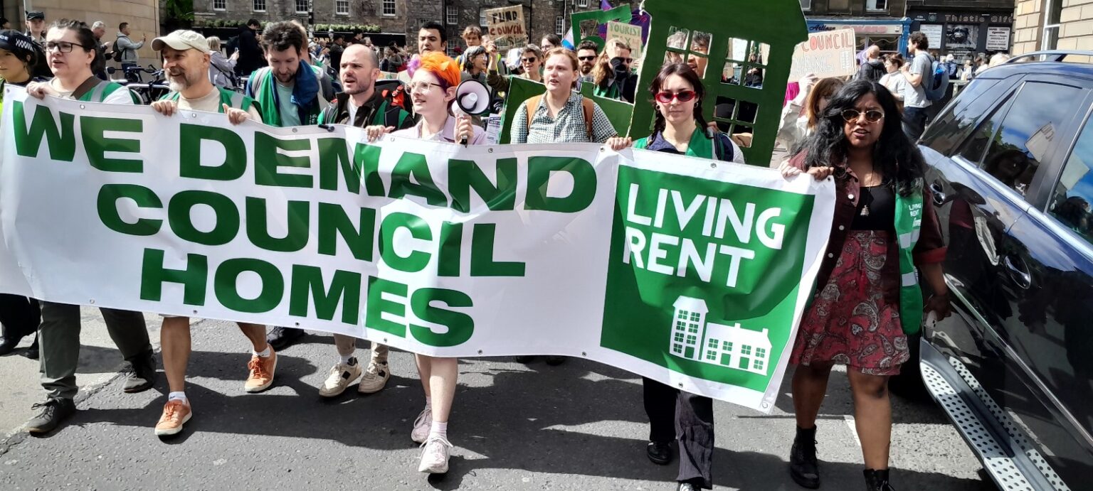 Attivisti di Living Rent Edinburgh marciano dietro uno striscione bianco con una scritta verde "We demand council homes"