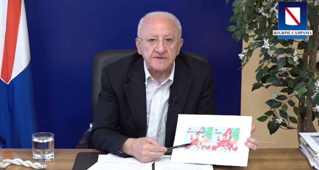 Il Presidente della Regione Campania Vincenzo De Luca, mentre parla dietro la scrivania del caso Gergiev. Su tavolo c'è un telefono e un bicchiere d'acqua alla sua destra; sullo sfondo la bandiera della Regione. Alla destra, sullo sfondo, uno pianta. De Luca parla tenendo in mano una penna, nell'altra ha una piantina che mostra i confini della Nato al momento della sua fondazione, e nel 2022.