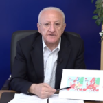 Il Presidente della Regione Campania Vincenzo De Luca, mentre parla dietro la scrivania del caso Gergiev. Su tavolo c'è un telefono e un bicchiere d'acqua alla sua destra; sullo sfondo la bandiera della Regione. Alla destra, sullo sfondo, uno pianta. De Luca parla tenendo in mano una penna, nell'altra ha una piantina che mostra i confini della Nato al momento della sua fondazione, e nel 2022.