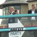 Nigel Farage con un megafono in mano accanto a Nathan Gill. I due stanno arrigando la folla dalla cima di un autobus a due piani con la parte sopra aperta. L'autobus ha i colori e il logo del "Brexit Party"