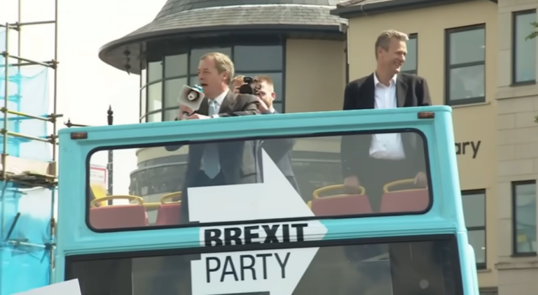 Nigel Farage con un megafono in mano accanto a Nathan Gill. I due stanno arrigando la folla dalla cima di un autobus a due piani con la parte sopra aperta. L'autobus ha i colori e il logo del "Brexit Party"