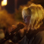 Diana Loginova di profilo davanti al microfono, mentre tiene in mano uno smartphone e si esibisce in pubblico